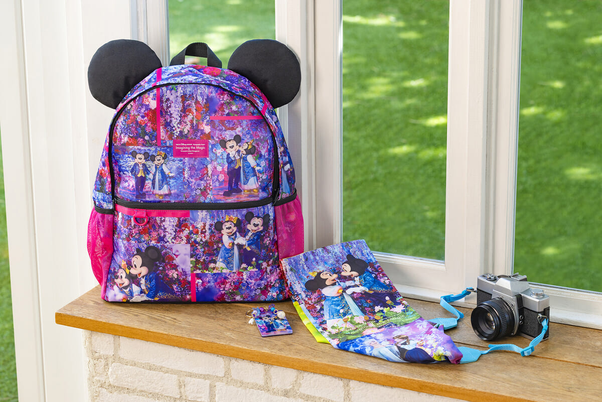 「ミッキーマウス」の耳が付いたアイコニックなリュックサックと、鮮やかなアートワークが目を引くカメラストラップ、レジャーシートが窓辺に並ぶイメージ