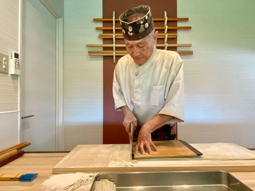 白い調理着を着た蕎麦職人が、打ち台の上で手打ち蕎麦を切っている作業風景。左手に駒板、右手に包丁を持ち、薄くたたんだ生地を均一な細さに仕上げている。手前には切りそろえた麺が並び、落ち着いた店内で名物の手打ち蕎麦を丁寧に作る職人技を伝える構図である。
