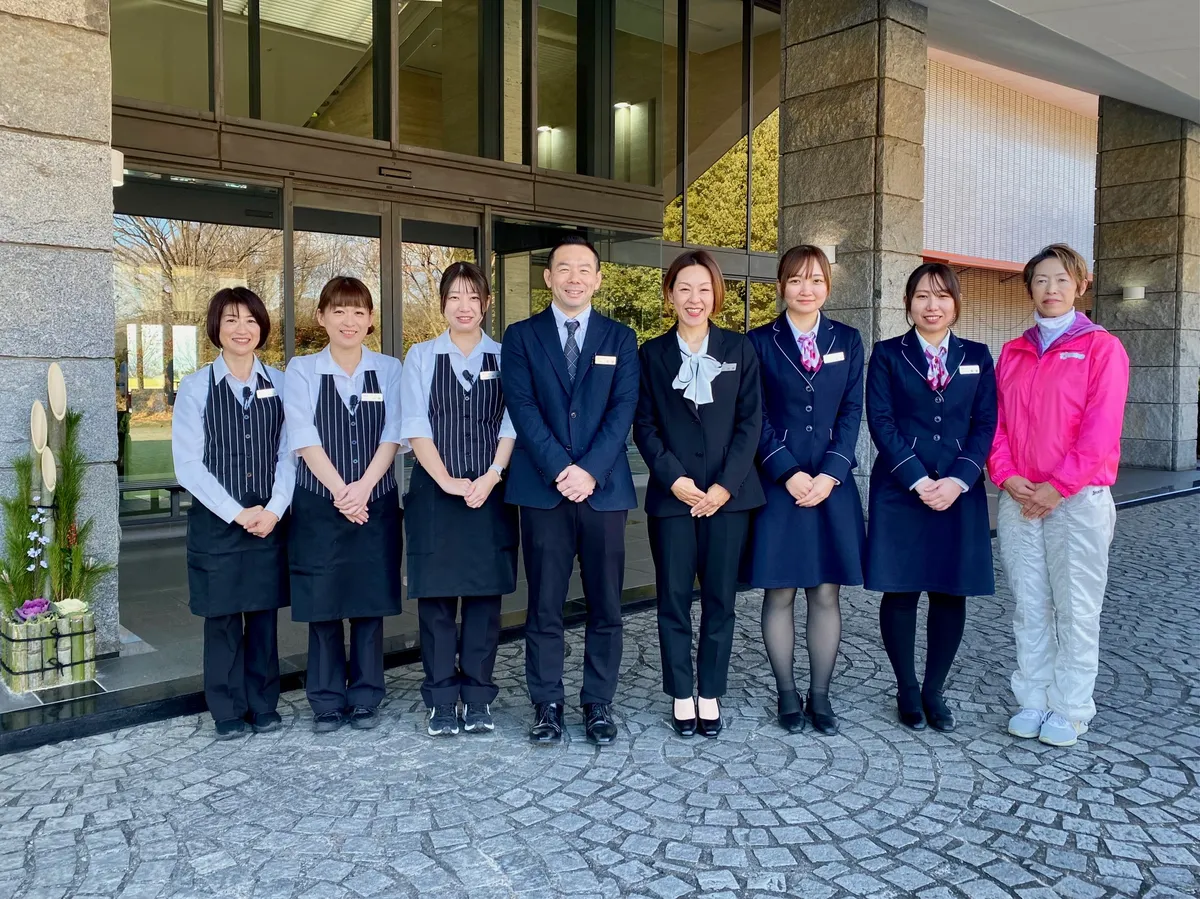 施設玄関前で8人のスタッフが横一列に並ぶ集合写真。飲食担当と思われるエプロン姿、スーツ姿の管理職やフロント担当、スポーツスタッフ風の制服姿がそろい、石造りのエントランスには門松も飾られている。ゴルフ場全体で来場者を迎える接客体制と一体感を示す記念的な構図である。