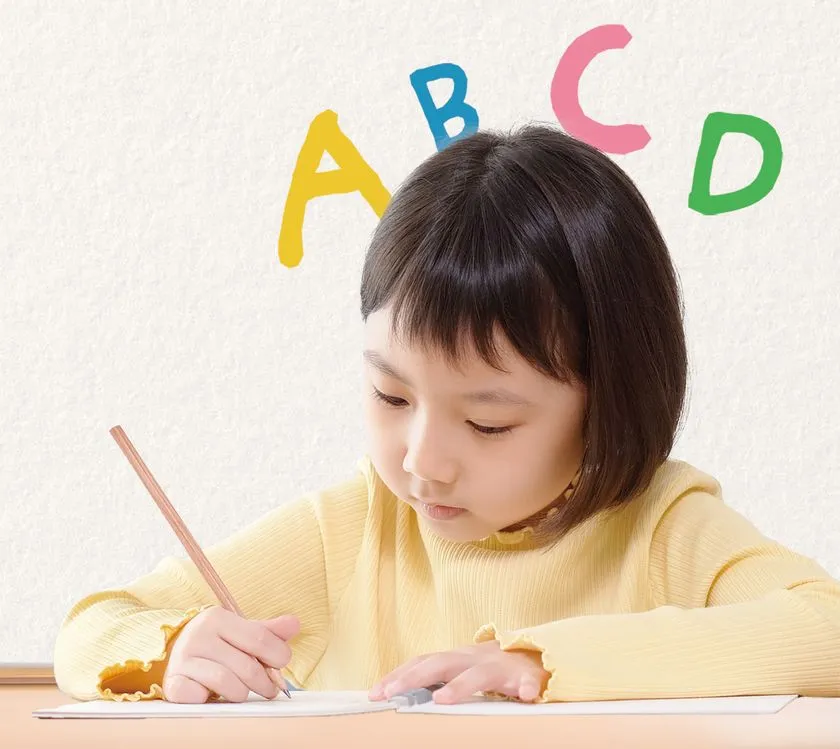 黄色いニットを着た幼児〜低学年の女の子が、机の前で鉛筆を握りノートに向かっている。頭上には「A・B・C・D」のカラフルなアルファベットが舞い、英語学習への第一歩を踏み出す子どもの姿を明るく表現。幼児・小学生向け英語教育のイメージに適した一枚である。