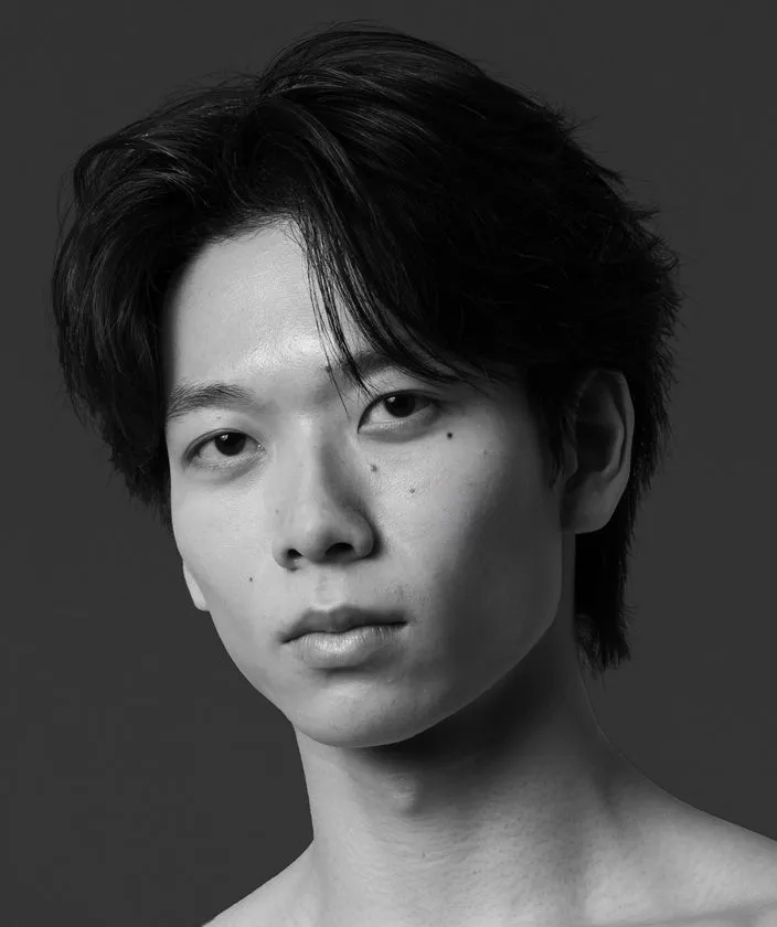 グレー背景の前で静かに視線を向ける若い男性のモノクロポートレート。黒髪を後ろにまとめ、上半身裸のシンプルな構図によって、引き締まった身体のラインと端正な表情が際立っている。ダンサーらしい繊細さと凛とした存在感が伝わる、舞台人の公式プロフィール写真にふさわしい一枚である。