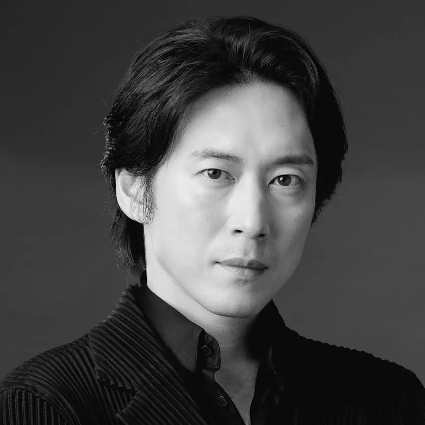 モノクロのスタジオポートレート。グレーの無地背景の前で、黒髪ストレートの女性がやわらかな微笑みを見せている。肩を見せた衣装に繊細なネックレスと小粒のイヤリングを合わせ、上品で洗練された印象を演出。舞台作品に関わる人物の気品と存在感を静かに伝える公式イメージ写真である。