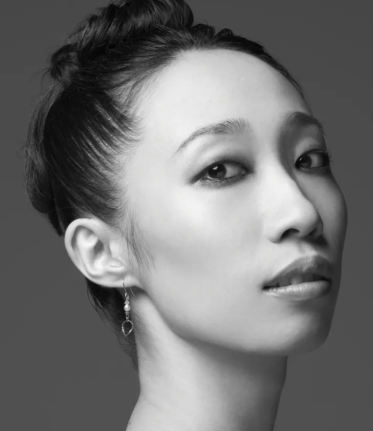 モノクロのスタジオポートレート。黒髪をタイトなシニョンにまとめた若い女性が、右手を頬に添えながら穏やかな微笑みを浮かべている。柔らかな光が顔立ちの陰影を美しく際立たせ、静かな気品と舞台人らしい洗練を感じさせる、公式プロフィール写真としてふさわしい一枚である。