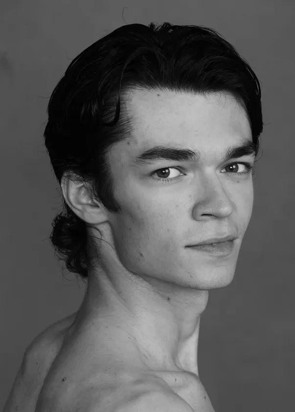 モノクロのスタジオポートレート。グレーの無地背景の前で、黒髪ストレートの女性がやわらかな微笑みを見せている。肩を見せた衣装に繊細なネックレスと小粒のイヤリングを合わせ、上品で洗練された印象を演出。舞台作品に関わる人物の気品と存在感を静かに伝える公式イメージ写真である。