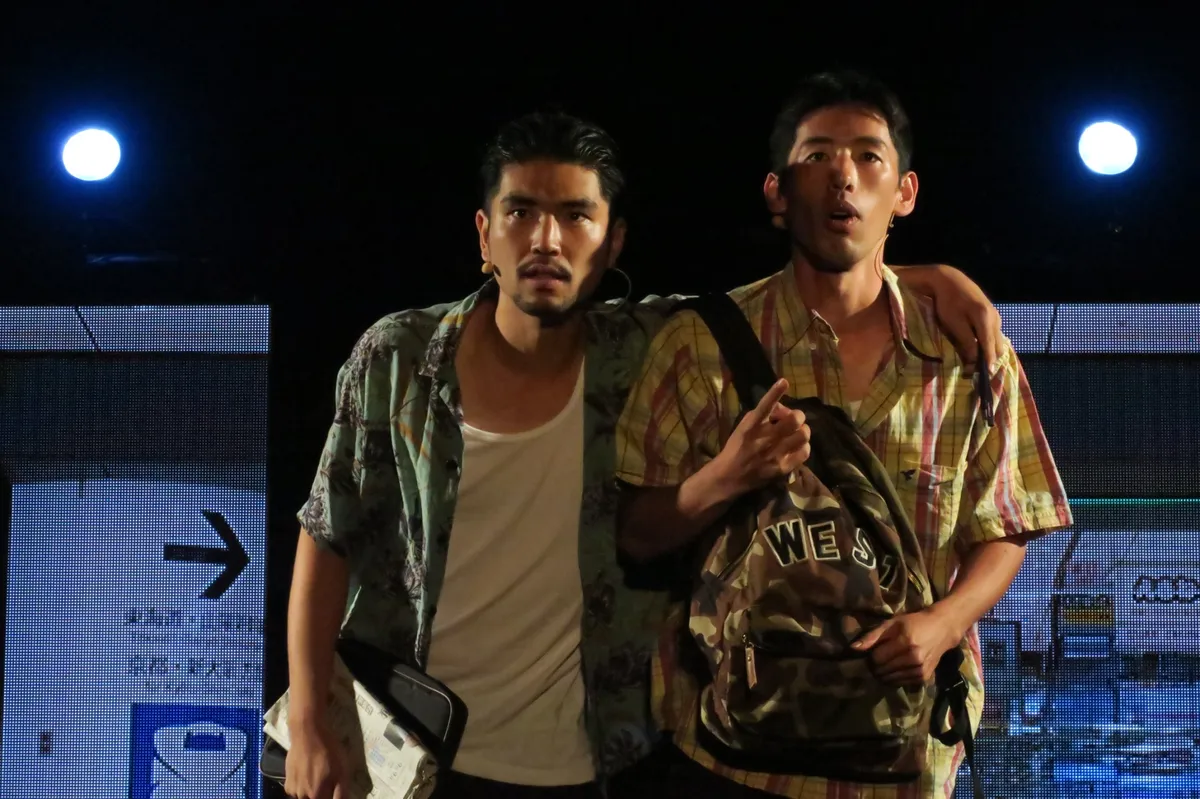 舞台上で肩を組む男性2人の本番シーン。空港セット背景にスポットライト、旅行バッグを手にした現代服の俳優2人が驚いた表情で上方を見上げている