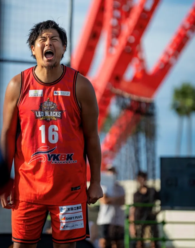 LEO BLACKS SAGA背番号16番の小堺翼が屋外コートで雄叫びを上げるプレー写真。背景に赤いクレーン構造物とヤシの木が写る