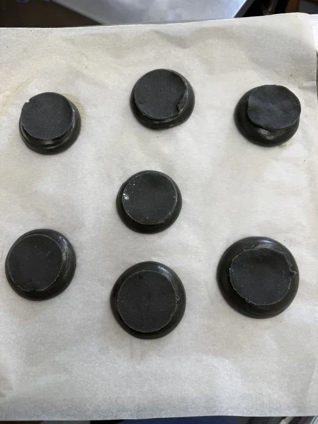 クリームを絞る前の黒いシュークリーム生地7個をクッキングシート上に並べた製造工程カット。墨色の均一な丸型生地が焼き上がった状態を真上から撮影