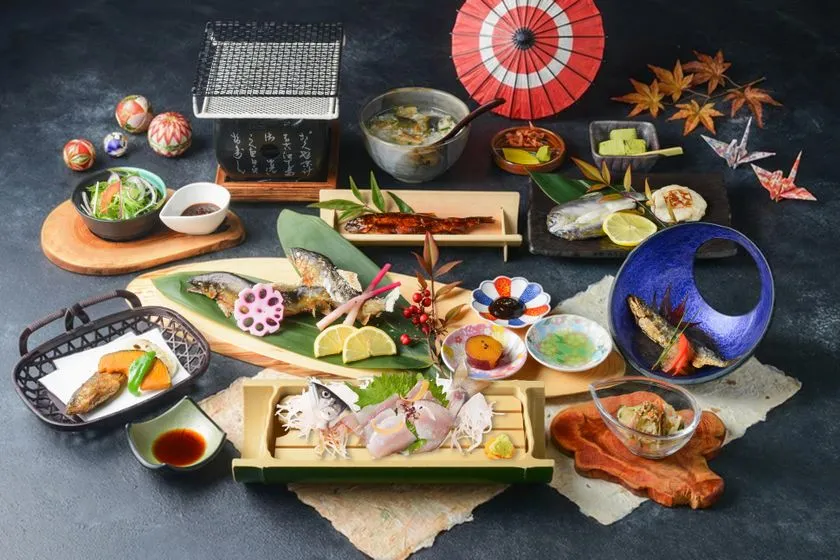 天然鮎の塩焼き・刺身・唐揚げ・お椀などを含む鮎料理フルコースの料理全体俯瞰写真。赤い和傘や紅葉の小道具とともに複数の器が並ぶ