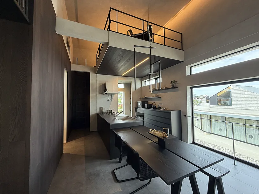 モデルハウス内部のダイニングキッチン実写。黒系アイランドキッチンとダイニングテーブル、鉄骨手すりのロフトスペースを上部に持つ吹き抜け空間
