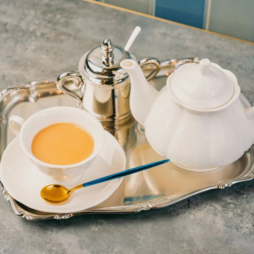 シルバートレーに白磁のティーポットとカップ・シュガーポット・ゴールドブルースプーンを並べたティーセット一式。紅茶またはミルクティーが注がれたカップが中央に配置