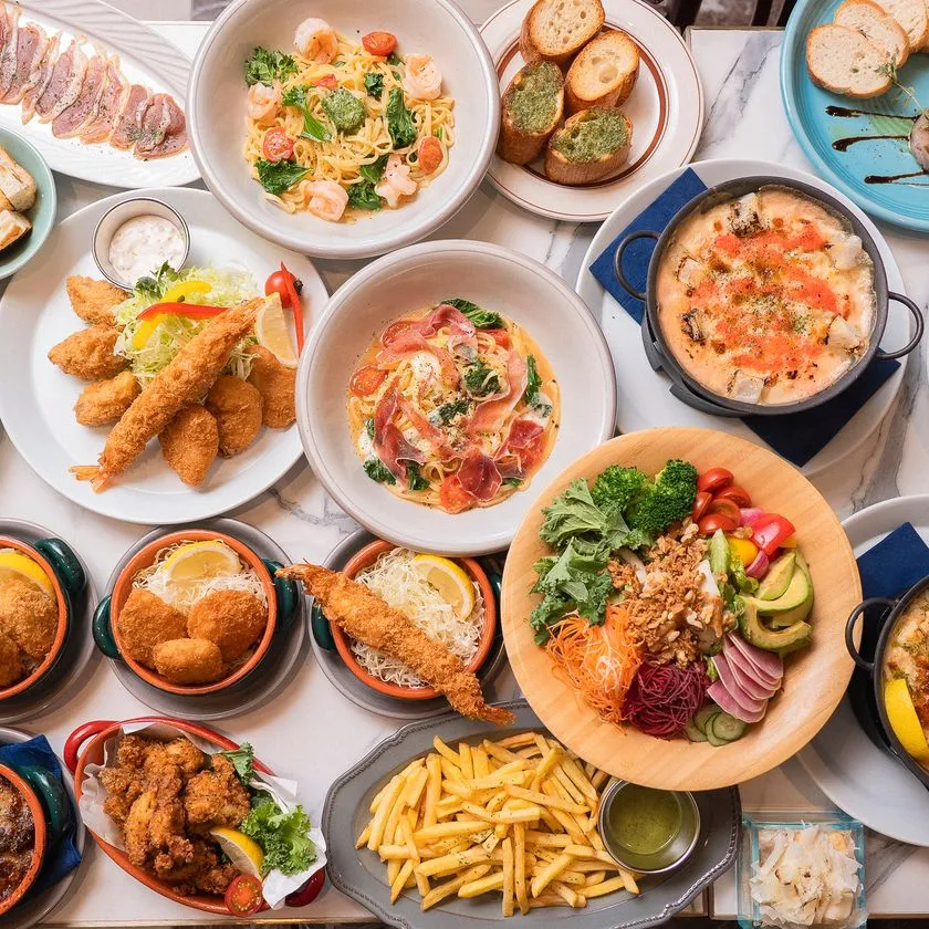 マルブルテーブルに並んだ多品目の料理フラットレイ。エビフライ・海老のパスタ・生ハムパスタ・フリット・フライドポテト・サラダボウル・グラタンなど10皿以上が俯瞰で写る