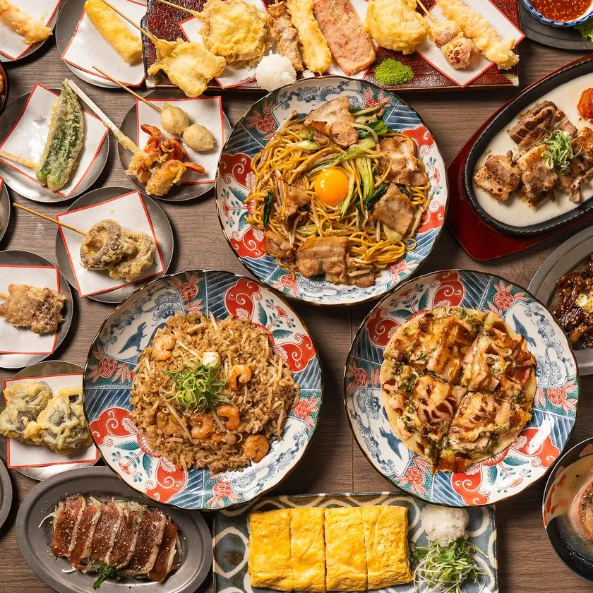 大衆酒場のコース料理を上から撮影した俯瞰写真。天ぷら串・焼きそば・海老チャーハン・お好み焼き・玉子焼きなど10皿以上が食卓に並ぶ豪勢な盛り合わせ。