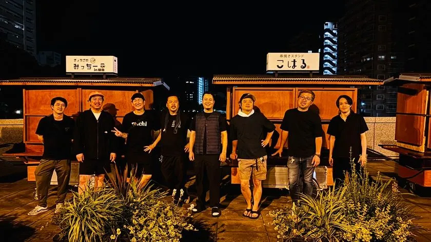 夜の甲突川沿いに並ぶリヤカー屋台「みっちー亭in鹿島」「串焼きスタンドこはる」の前で、黒Tシャツ姿の男性スタッフ8名が横一列に並んで記念撮影している集合写真