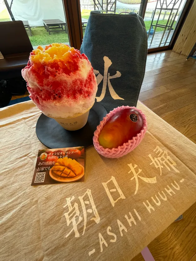 台湾人気店・朝日夫婦のかき氷とチャンピオンマンゴー1玉をテーブルに並べた物撮り。赤とオレンジのグラデーションかき氷と朝日夫婦ロゴ入りクロスが写る