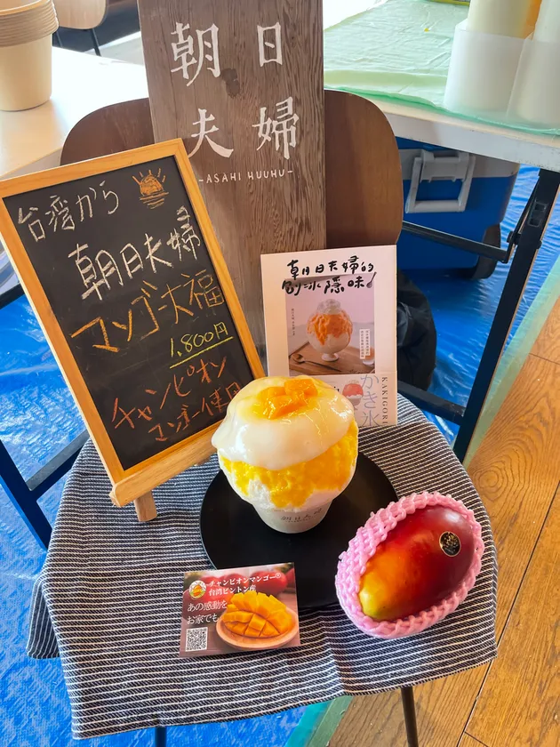 朝日夫婦ブースの販売台に並べたマンゴー大福かき氷1,800円のメニュー黒板・かき氷・チャンピオンマンゴー実物・QRコードカードのセット展示ショット