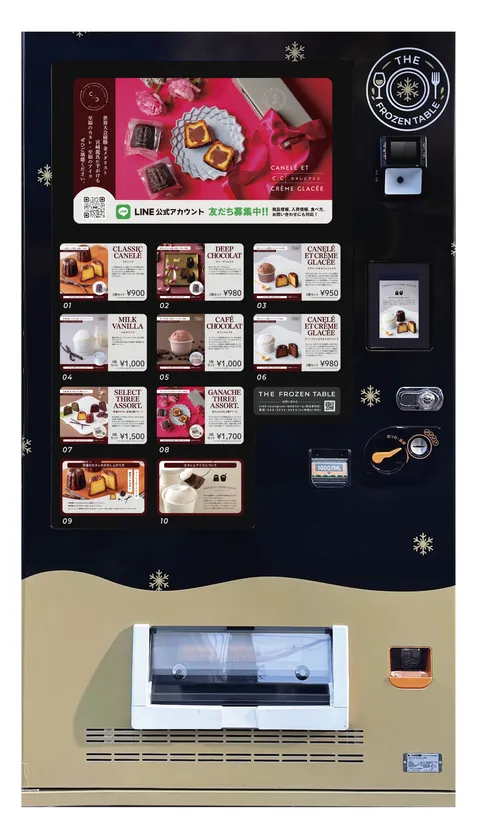 THE FROZEN TABLEの冷凍自動販売機全景。カヌレ・アイス・アソートなど10種のメニューパネルと各価格（900円〜1,700円）が表示され、LINEアカウントの案内も掲示されている