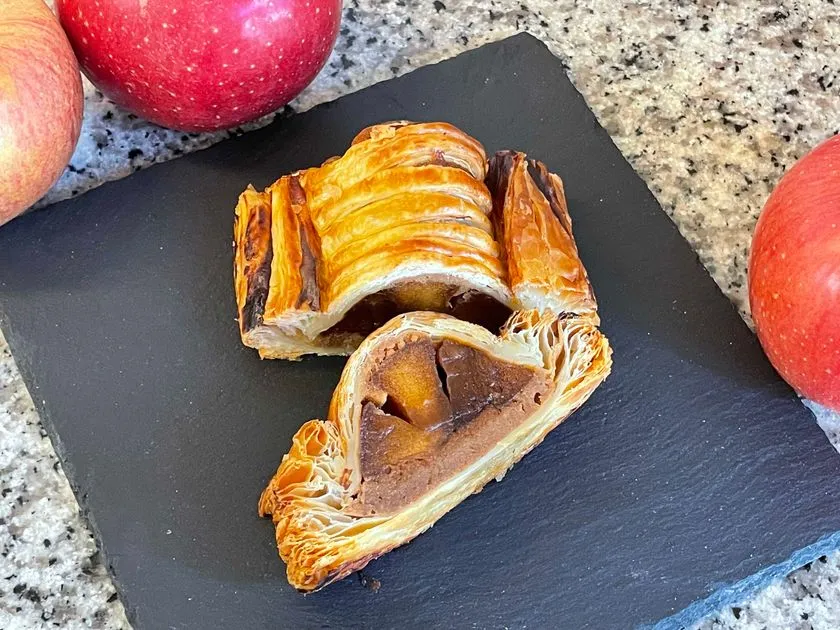 パイ生地を割った断面にりんごとチョコレートクリームの詰め物が見えるアップルパイ系焼き菓子の商品ショット、スレートプレートに赤いりんごを添えて撮影