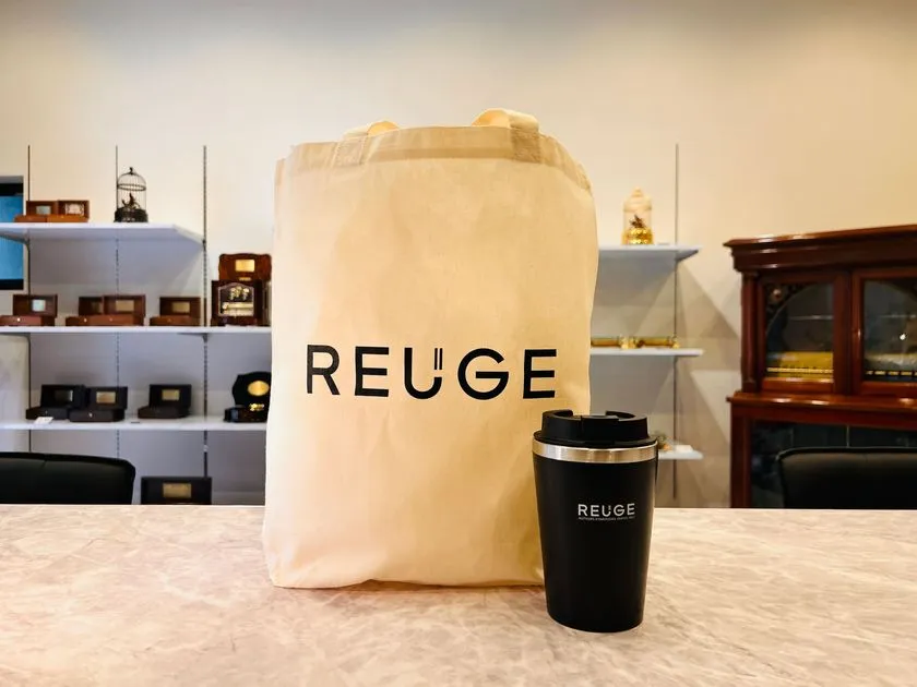 REUGEロゴ入りキャンバストートバッグと黒いステンレスタンブラーを店頭カウンターに並べたノベルティ2点セット。背景の棚にはREUGEオルゴール複数台が陳列されている。