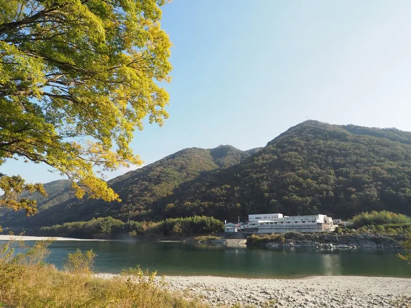 吉野川沿いの河川敷と山を背景に、株式会社オカベの白い工場建物を望む自然環境の外観ショット