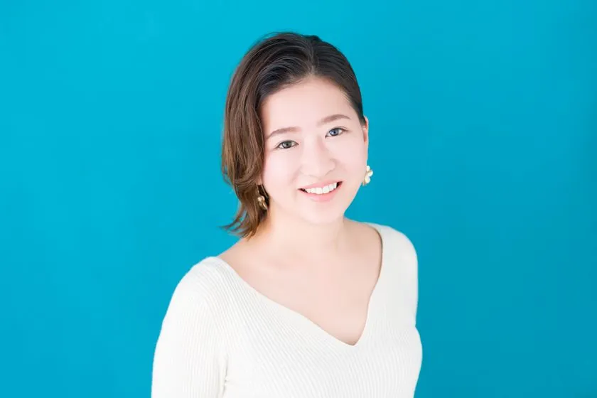 水色背景に白ニットを着た短髪女性のプロフィール写真。正面向きの上半身ポートレート