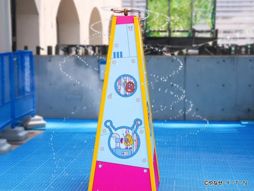 バイキンメカをモチーフにしたピンク・黄色の三角錐型ウォータースプレー装置が水を四方に噴射している様子。青いプールエリアの床面上に設置されている。
