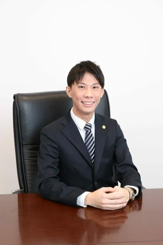 黒スーツ・ストライプネクタイ姿の男性弁護士がレザーチェアに座り両手を重ねて正面を向いた公式プロフィール写真、胸元に弁護士バッジあり