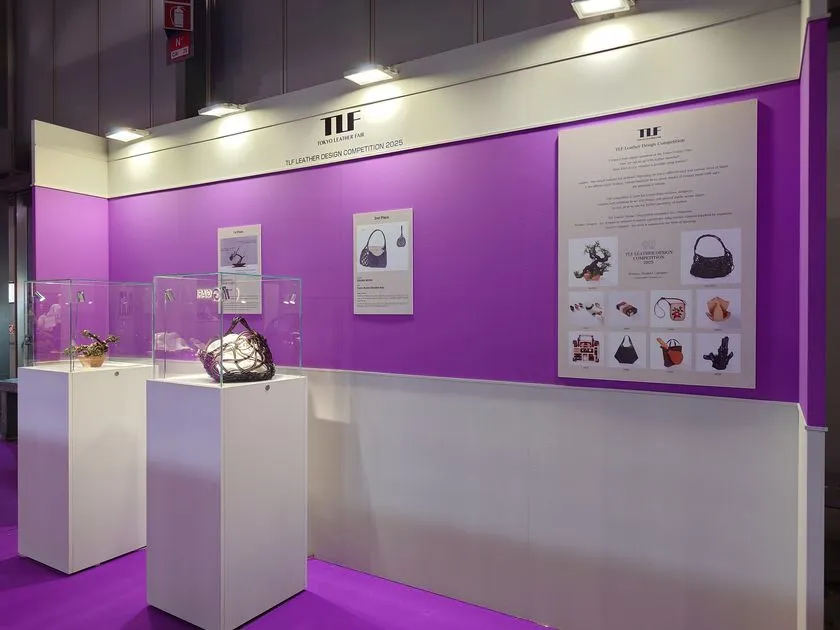 過去回のTLFレザーデザインコンペ展示ブースの全景写真。紫色の壁面パネルに受賞作品パネルを掲示し、白い台座上にガラスケース入り受賞革製品バッグ2点を展示
