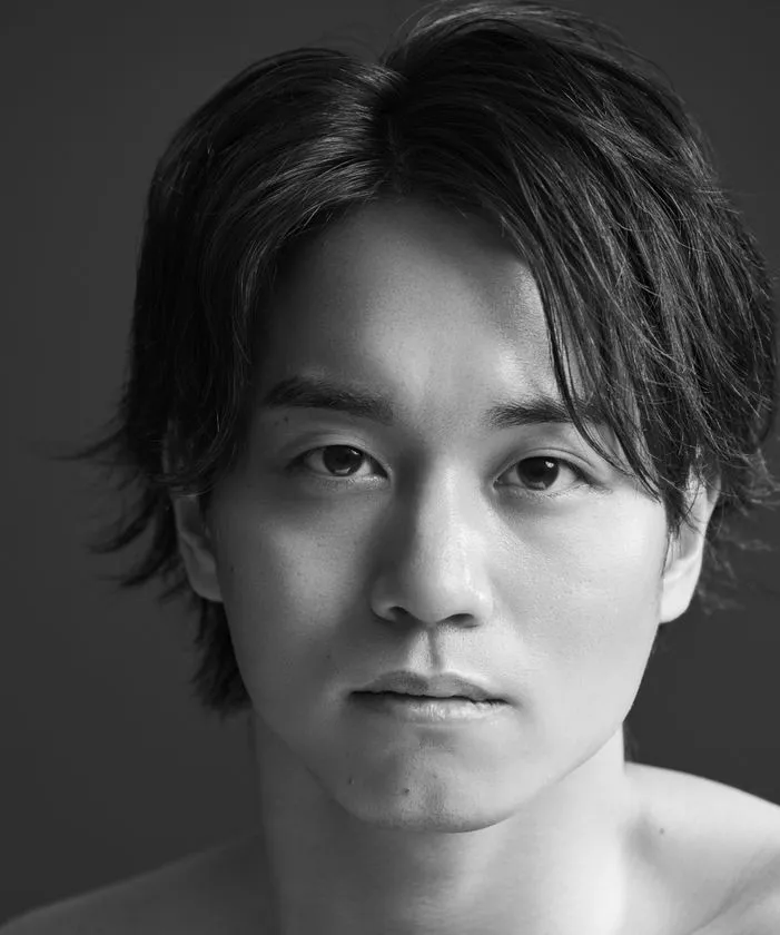 K-BALLET TOKYOの男性ダンサーとみられる人物のモノクロ公式プロフィール写真。中長めの黒髪と上半身が映るグレー背景のクローズアップ構図で真剣な表情。