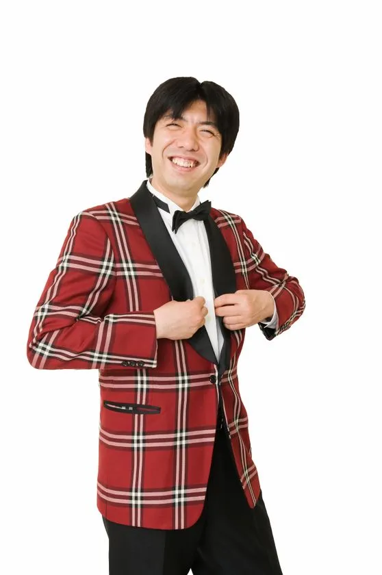 赤チェック柄タキシードジャケットに蝶ネクタイを着用した男性芸人の宣材写真。白背景で笑顔のポーズ。