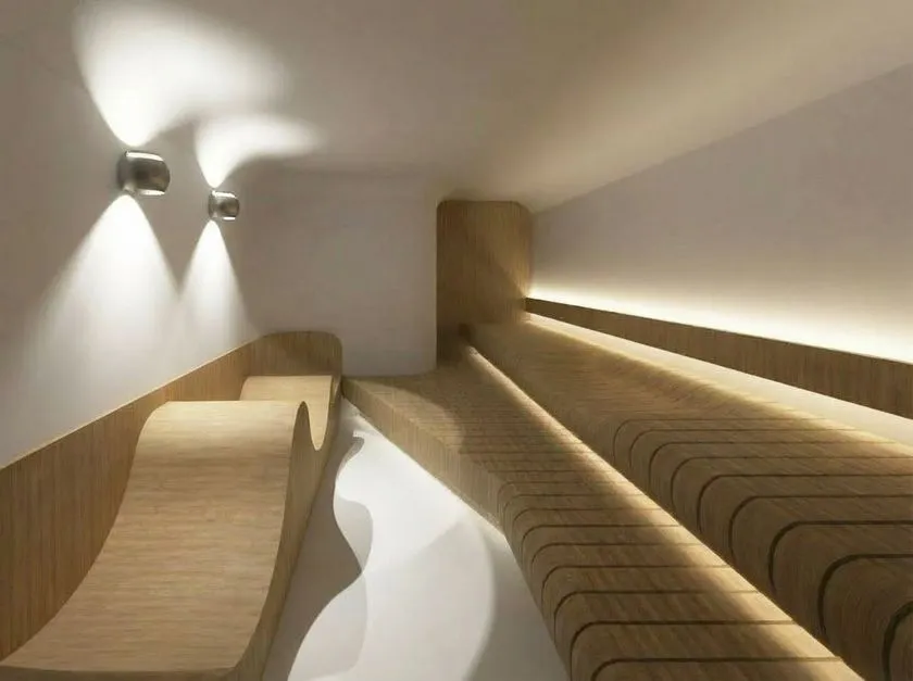 洞サウナ(DO SAUNA)の室内撮影写真。曲線状の木製ベンチが2段に並ぶサウナ室内に間接照明と壁付きスポットライトが灯り、洞窟をイメージした白壁と木材の内装デザインが確認できる。