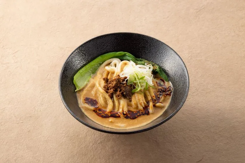黒陶器の丼に盛られたグルテンフリー玄米うどんの担々麺風メニュー。太麺に濃厚スープ、肉味噌、青梗菜、白髪ねぎをトッピングしたメニュー写真