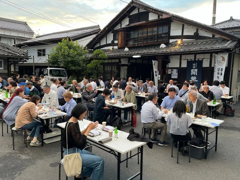 第一酒造の蔵元敷地内屋外会場に多数の参加者がテーブル席に着き、日本酒瓶を囲んで飲食している夕刻の「ひやガーデン」会場全景。白壁の蔵建物と「開華」のれんが背景