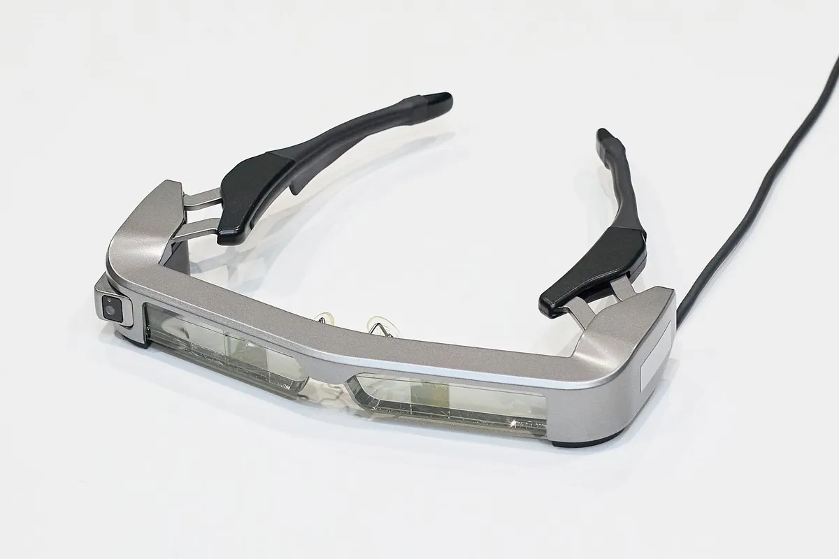 視覚障がい者向け道案内デバイスのスマートグラス本体。シルバーフレームにレンズユニットとカメラを内蔵し、黒いテンプル（つる）とケーブルが接続された斜め俯瞰の製品単体ショット。