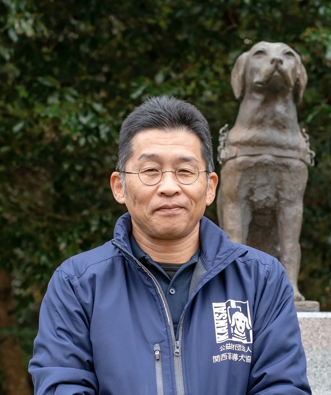 公益財団法人関西盲導犬協会のジャケットを着た男性スタッフが、盲導犬をかたどったブロンズ像を背景に正面を向いて立つポートレート写真