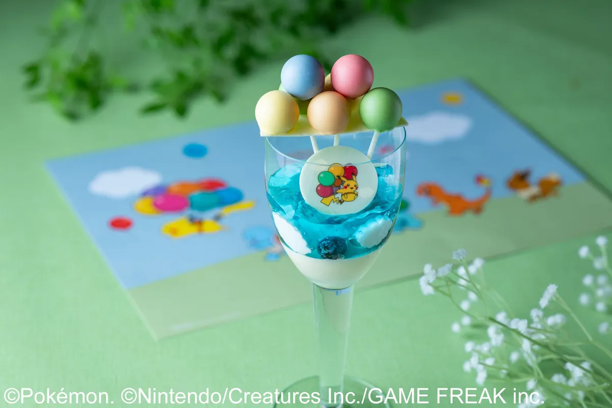 青いゼリーとクリームを重ねたグラスデザートの上部にパステル5色のケーキポップ球が串刺しで飾られ、ピカチュウとバルーン柄のチョコレートメダルが側面に添えられたポケモン30周年スイーツ