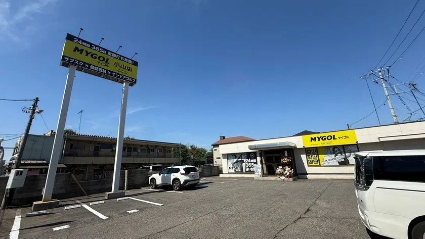 マイゴル小山店の駐車場と外観全景。24時間365日定額打ち放題を謳うポール看板と店舗建物全体が写り、広い駐車スペースが確認できる