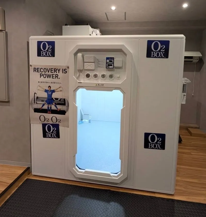 店内に設置された酸素カプセル「O2 BOX」の正面写真。白色の大型筐体にブルーライトが灯った入口扉があり、RECOVERY IS POWERのポスターと操作パネルが確認できる施設設備ショット
