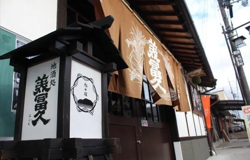 地酒処「兼冨久」の外観写真。水口宿の行灯看板と茶色地に兼冨久と染め抜かれた大暖簾が並ぶ酒蔵の正面