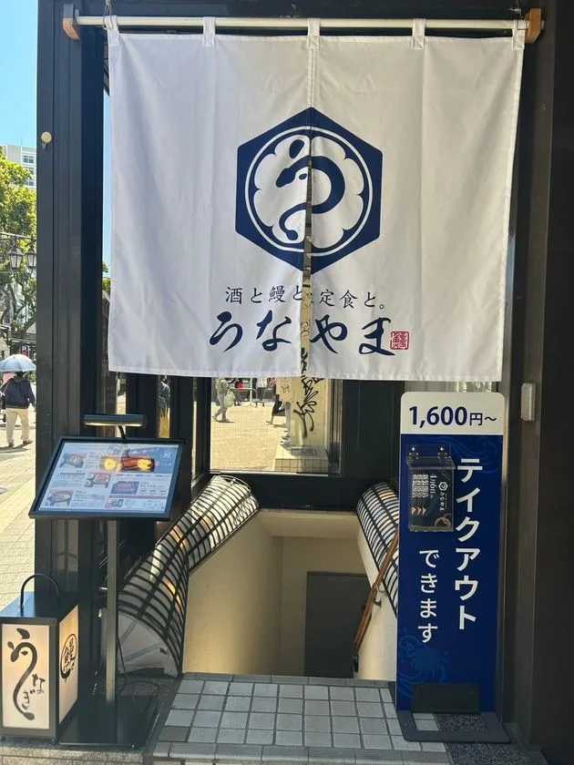 うなやまの店舗正面外観。白い暖簾に店名ロゴと酒と鰻と定食との文字、1600円〜のテイクアウト案内看板とメニューボードが確認できる