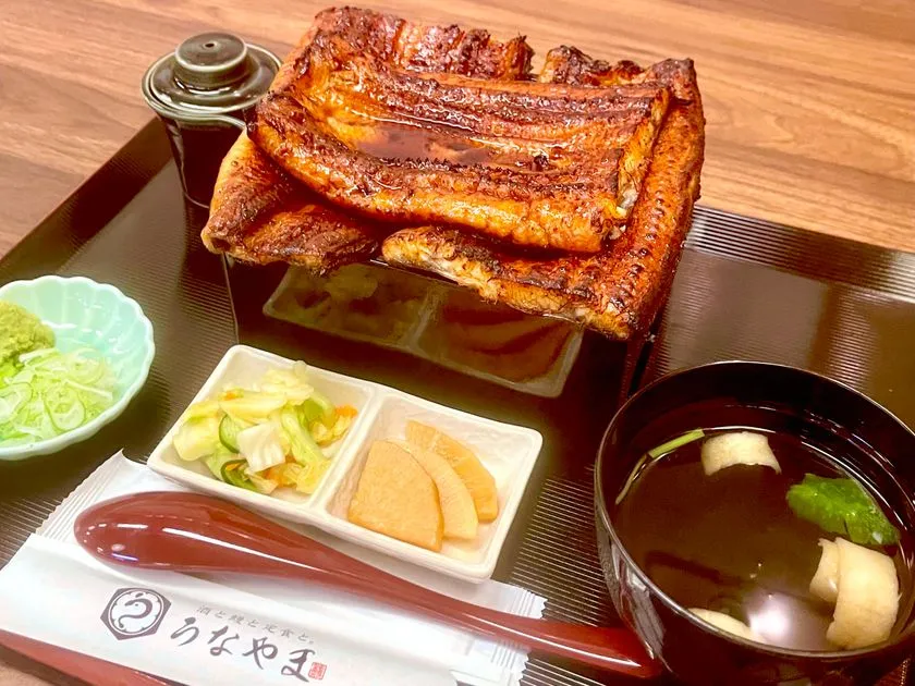 マウンテン重の斜め横アングル撮影。重箱から立体的に盛り上がった鰻の蒲焼き2匹分の厚みと高さが強調され、漬物・お吸い物・薬味の小皿とうなやまロゴ入り箸袋が添えられる