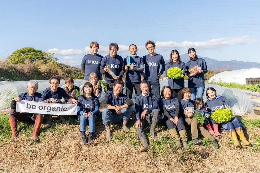 株式会社いかすのスタッフ約14名がbe organicバナーと野菜を持ち、農場ビニールハウスを背景に集合した屋外チーム写真