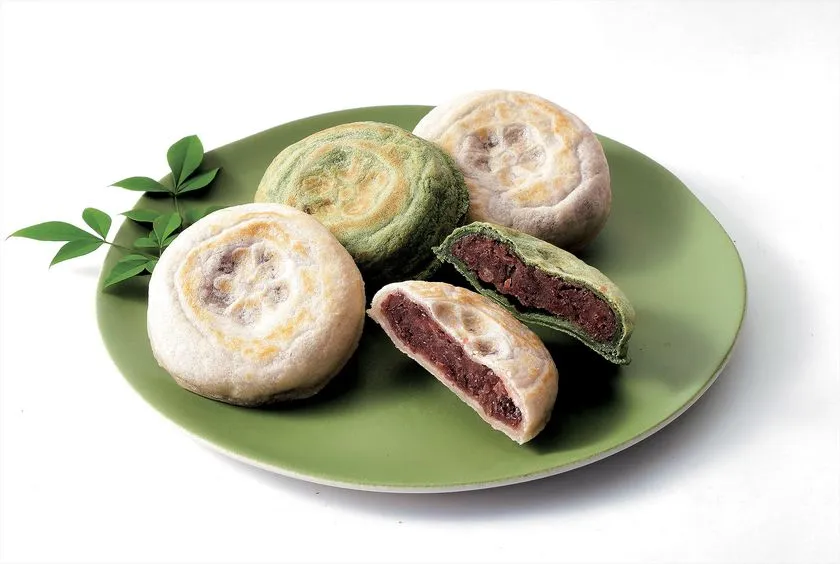 緑皿に並んだ焼き饅頭3個と断面カット1個の商品写真。白皮・抹茶皮の2種があり、断面にこしあん入りの饅頭の内側構造が確認できる