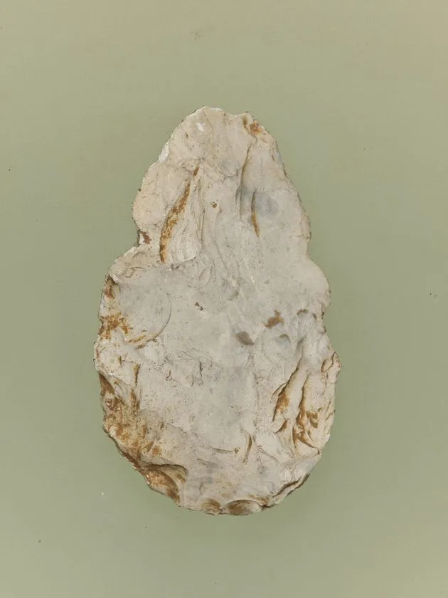 旧石器時代の握斧（ハンドアックス）または礫器を緑灰色背景で単体撮影。白色チャート質の扁平な楕円形石器で、表面に複数の剥離痕と錆色の鉄分変色が見られる