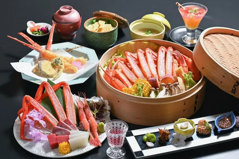 蟹を主役にした旅館の夕食膳全景。蒸籠に盛られた蟹脚の蒸し料理、蟹刺し、海老天ぷら、小鉢5種、スープ、フルーツカクテルが黒いテーブルに並ぶコース料理の全品配膳ショット