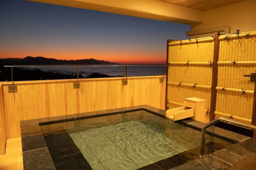 夕暮れ時の夕日ヶ浦海岸を望む客室露天風呂。石張りの浴槽と木製の目隠し竹垣、オレンジ色に染まる水平線と海が一望できる屋外温泉浴槽のショット