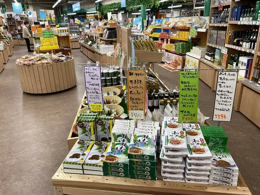 道の駅しもにた店内の物産コーナー全景。下仁田ねぎみそ・ねぎ味噌・青唐辛子加工品など箱入り商品が木製台に積まれ、702円・648円・594円の値札と手書きPOPが付いた特産品売り場の様子