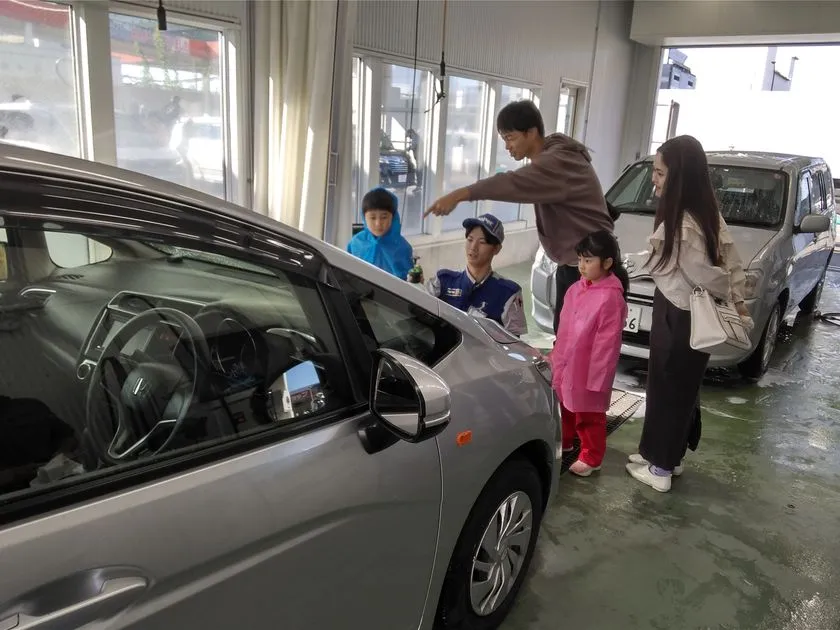 カーケアセンター内で整備士スタッフが親子連れに車のボディを指差しながら説明しており、子どもがピンクのレインコートを着て参加している様子