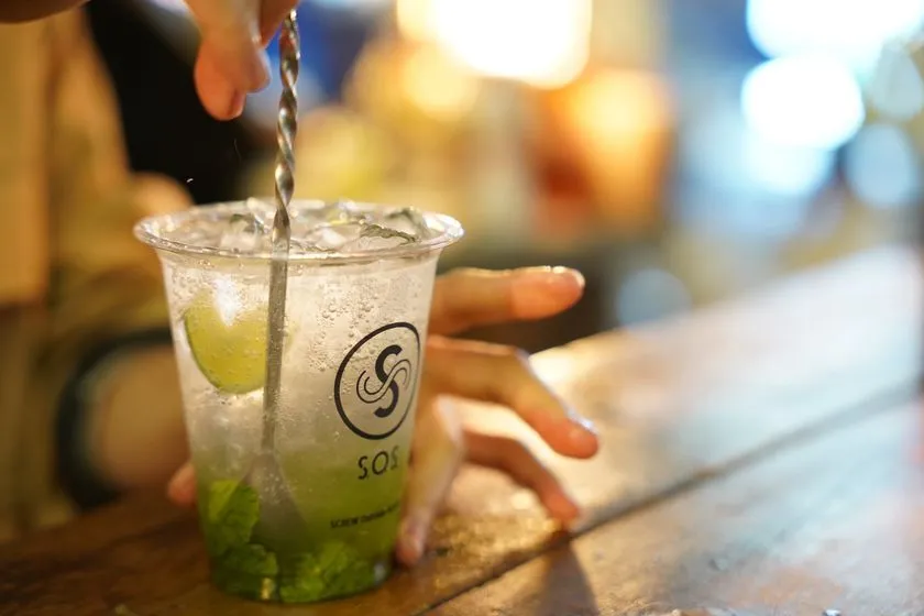 バーテンダーがSOSロゴ入りプラスチックカップに入ったライムとミントのカクテル（モヒート系）をステアしているアップショット