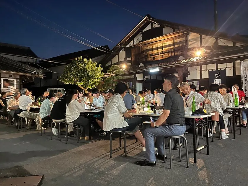夜間のひやガーデン開催時の屋外宴席風景。ライトアップされた酒蔵前で参加者が日本酒瓶を囲んでテーブルごとに着席し、夜の酒蔵ガーデンでの飲食シーンを写している
