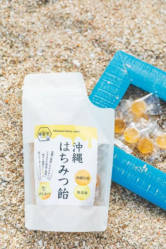 蜂蜜堂の沖縄はちみつ飴(Okinawa honey candy)スタンドパック正面と個包装キャンディが砂浜に置かれた商品写真、沖縄県産・無添加・生はちみつ表記あり